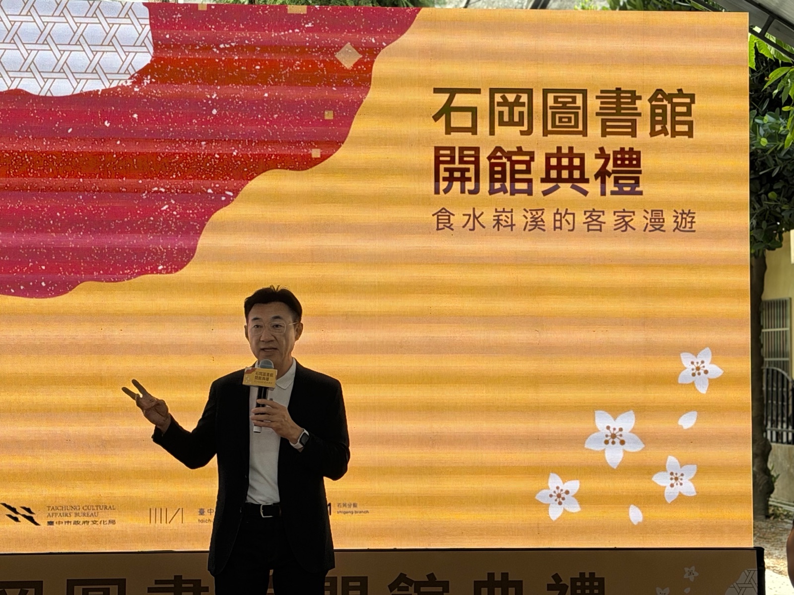 穿深色西裝外套的男子手持麥克風致詞，身後大螢幕寫著「石岡圖書館 開館典禮 食水嵙溪的客家漫遊」