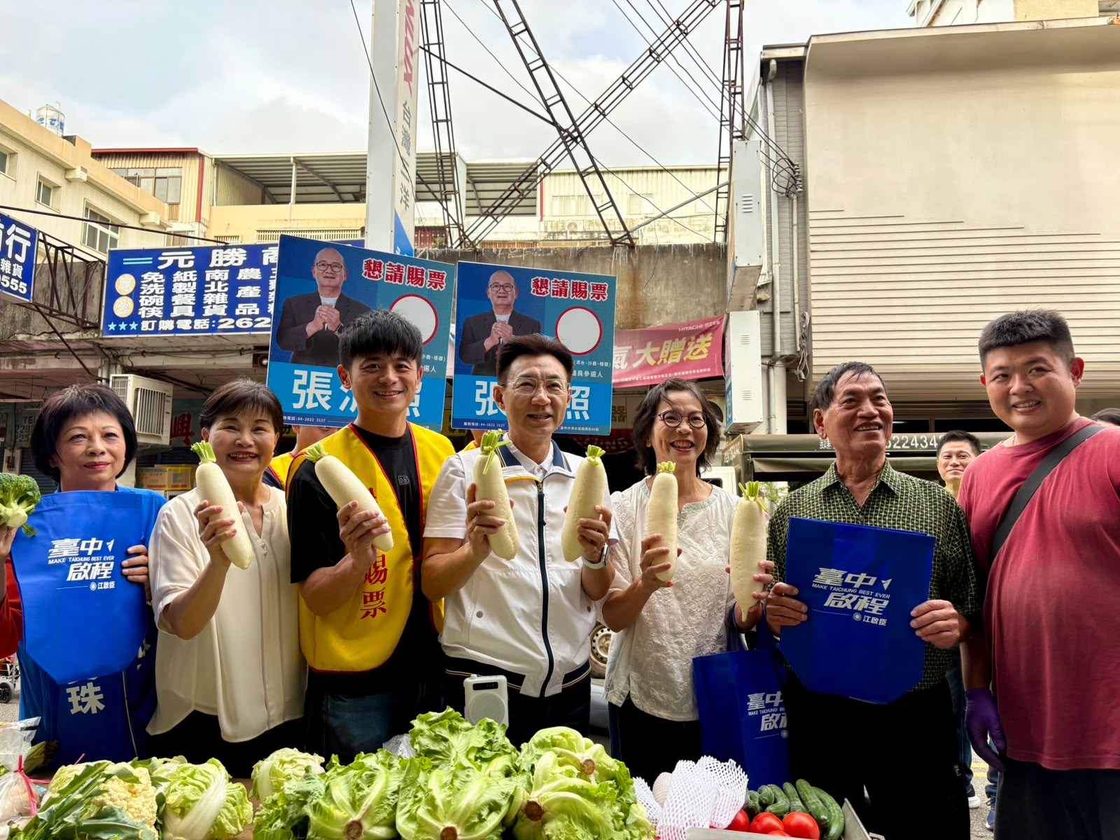 菜攤前，穿白色運動外套的男子與多位民眾手捧白蘿蔔合影，部分人拿著「臺中啟程 江啟臣」藍色提袋，後方立著「懇請賜票 張清照」看板