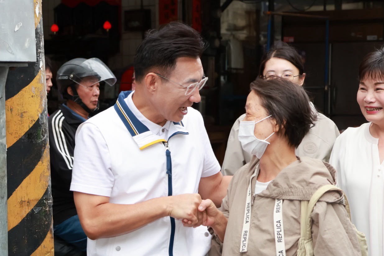 街邊穿白色運動外套的男子與一位戴口罩的市民女士握手，兩人笑著交談，旁邊另有兩位女士微笑