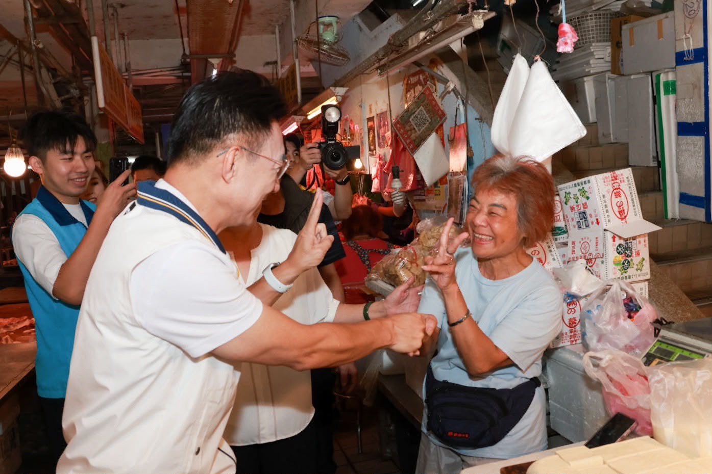 市場內穿白色運動外套的男子與一位穿淺藍上衣的攤商女士碰拳、比出手勢，後方穿藍背心的年輕人與多位民眾拿手機拍攝