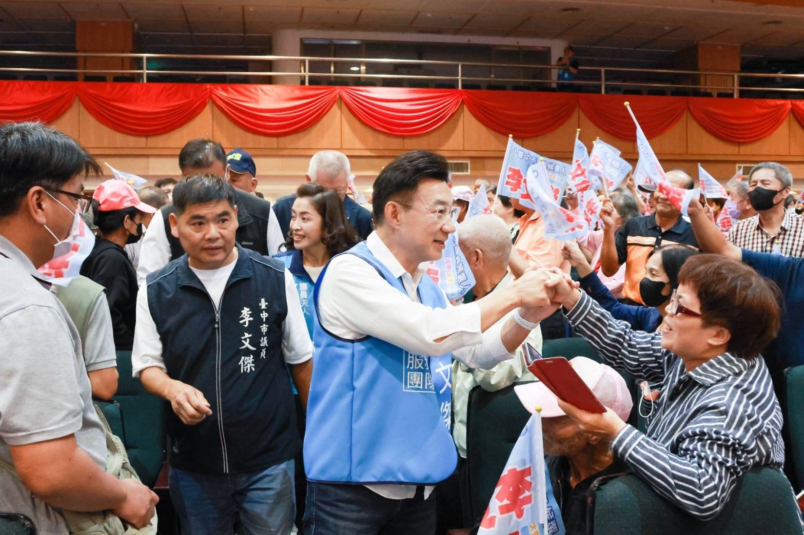 大甲會場走道上，穿「李文傑 服務團隊」藍背心的男子與臺下民眾握手，左側穿深色「臺中市議員 李文傑」背心的男子隨行，四周民眾揮舞「李文傑」小旗