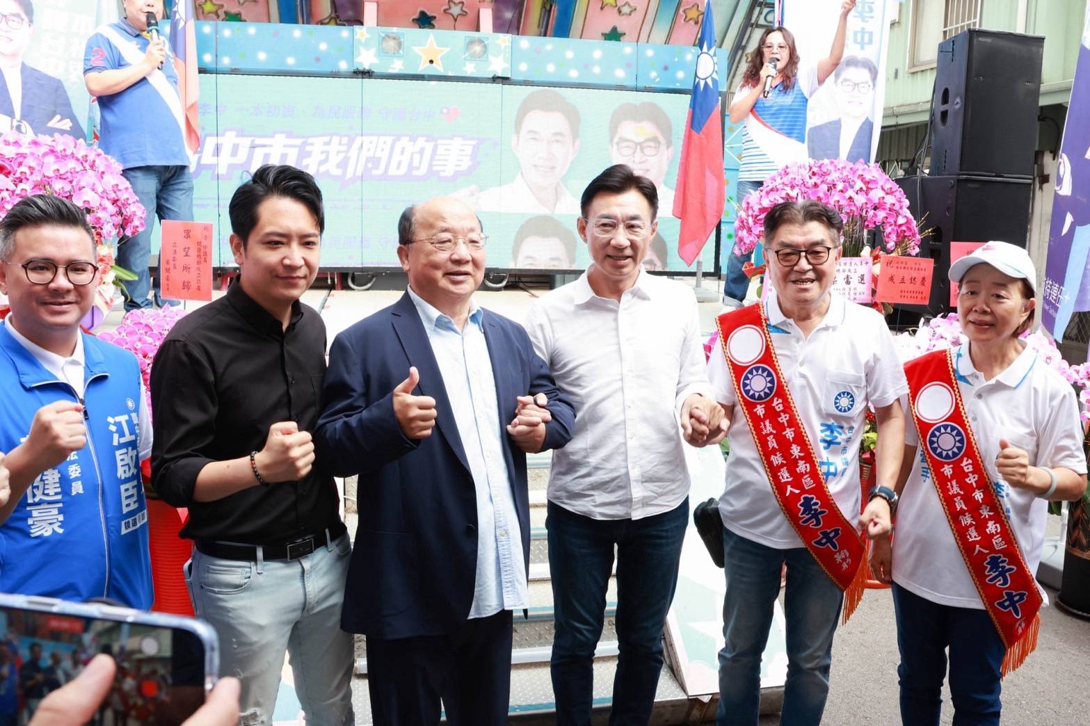 東區聯合競總成立大會，穿白襯衫的男子與穿深藍外套、比讚的長者及多位來賓合影，右側兩人披著紅色「市議員候選人 李中」肩帶，左側穿藍背心「立法委員 黃健豪」，背後螢幕寫著「臺中市我們的事」