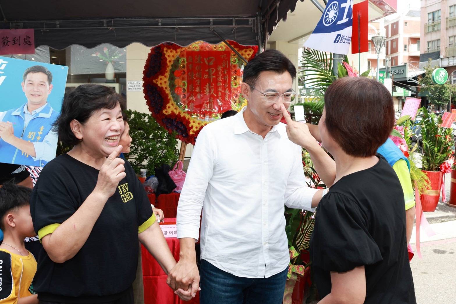 劉士州競選總部成立大會會場外，穿白襯衫的男子笑著讓一位市民女士伸手點他的鼻子，旁邊另一位女士也笑著比出手勢，後方是「劉士州」看板與賀牌