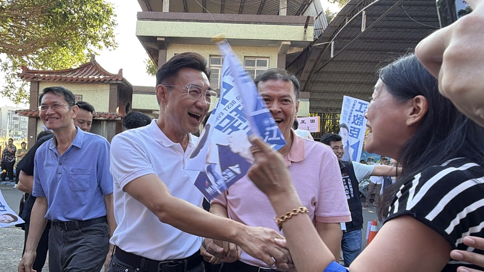 穿白色Polo衫的男子笑著與迎面的女士握手，手持「臺中啟程」小旗的鄉親簇擁在旁，後方立著「江啟臣」看板