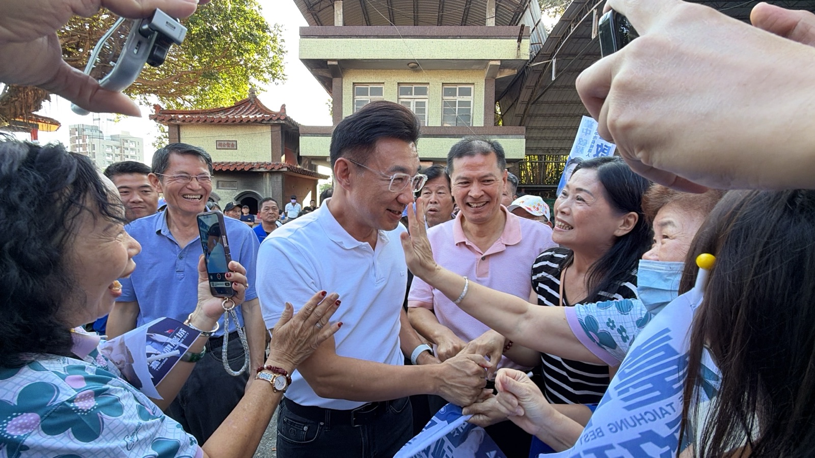 穿白色Polo衫的男子進場時被鄉親團團圍住，眾人伸手握手、舉起手機拍照，後方是廟宇與鄉間建築
