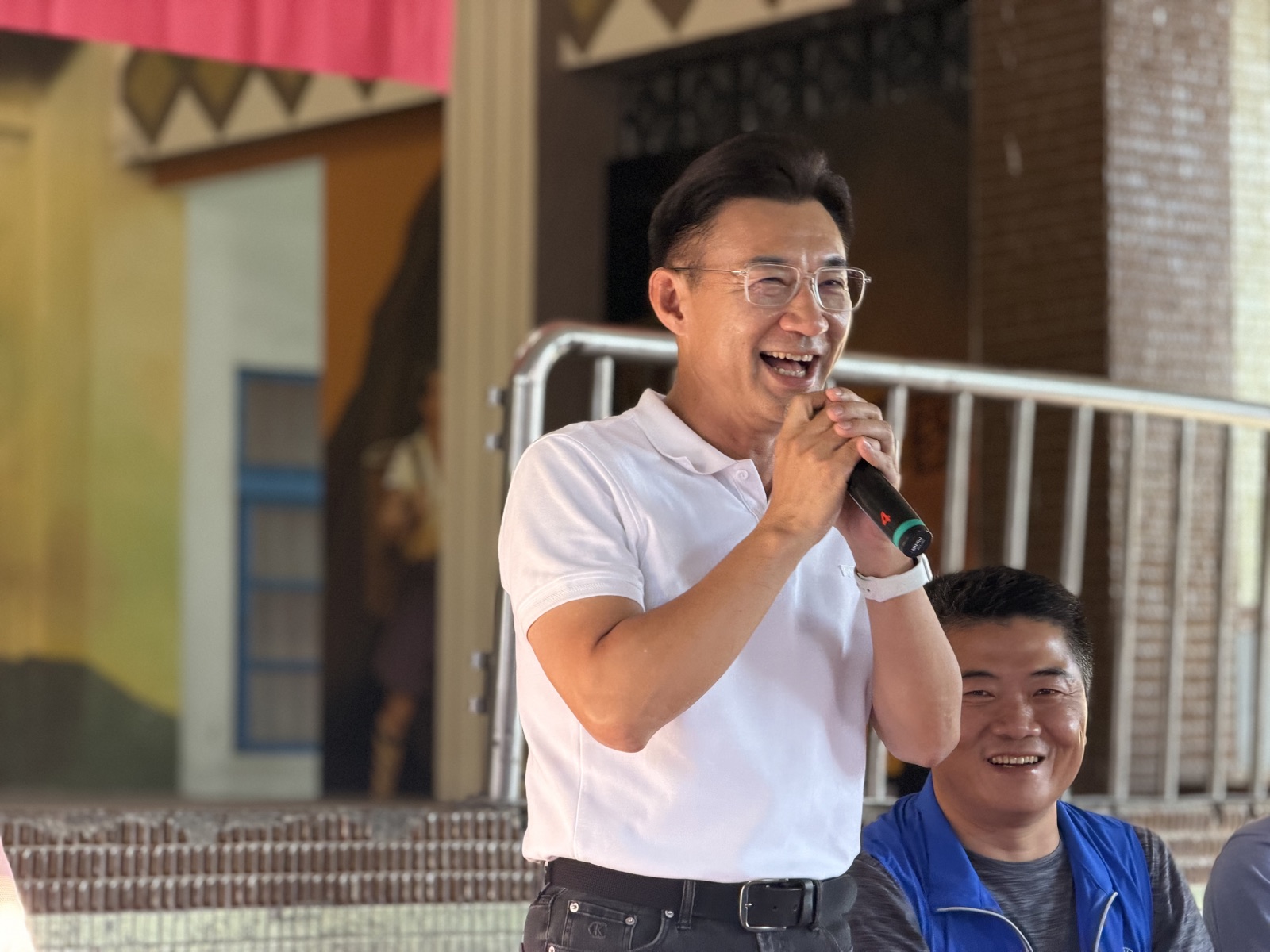 穿白色Polo衫的男子笑著雙手握麥克風發言，身後坐著穿藍色背心的男子，背景為廟埕的彩繪牆面