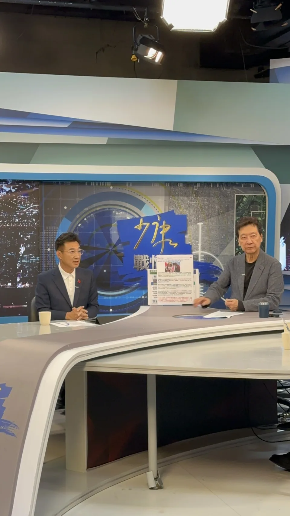 攝影棚內，穿深藍西裝、白色上衣的男子雙手交握坐在「戰情室」布景前的弧形桌後開口說話，右側穿灰色外套的主持人低頭看著桌上的資料，桌前放著白色馬克杯