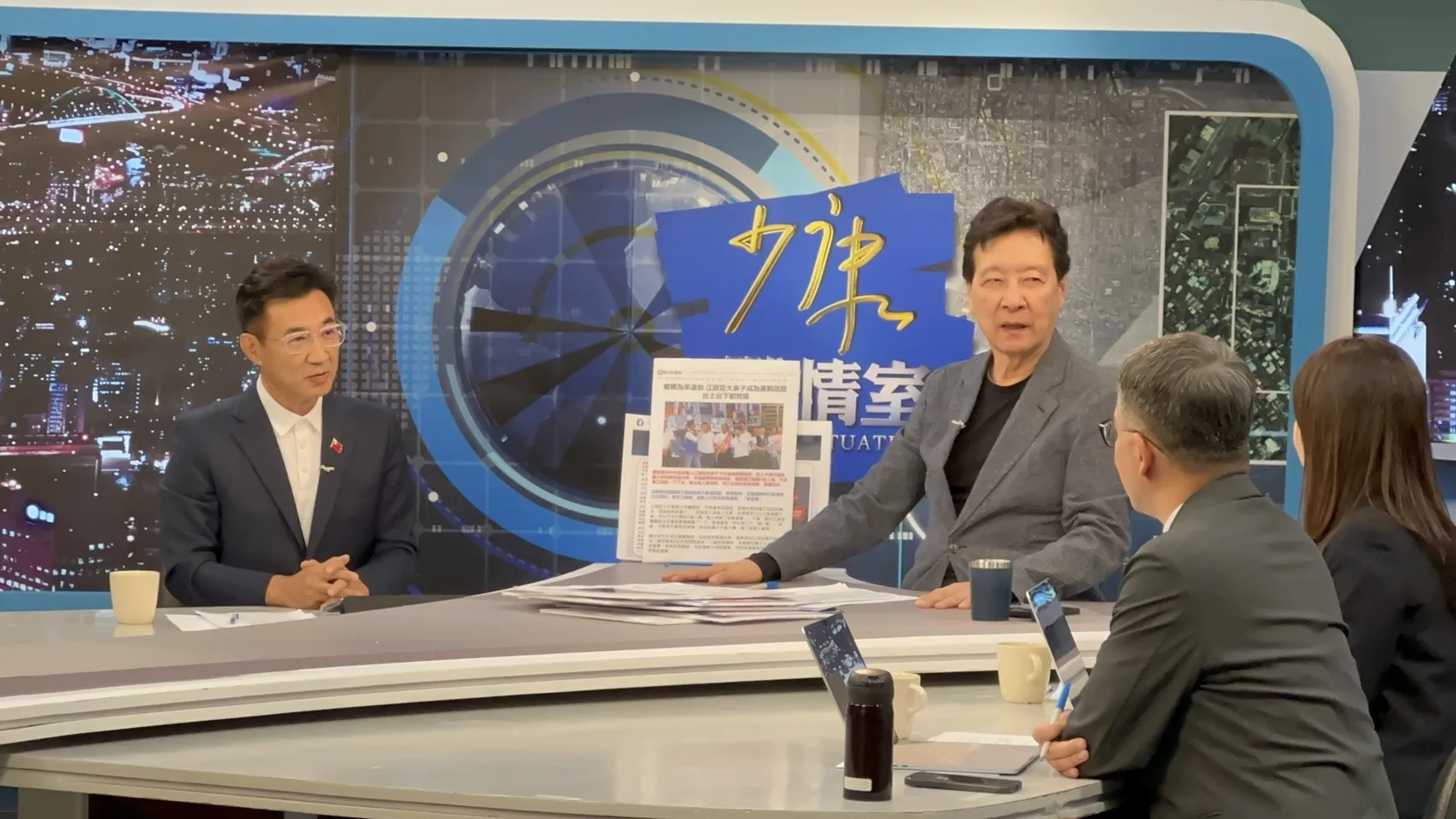 《少康戰情室》攝影棚內，穿深藍西裝、白色上衣的男子雙手交握坐在左側受訪，中間穿灰色外套的主持人伸手指向立在桌上的剪報，背景是藍色「戰情室」布景，右前方坐著兩位背對鏡頭的工作人員