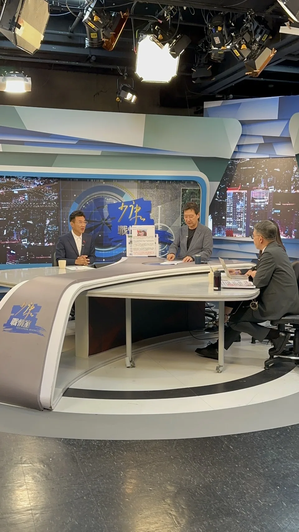 《少康戰情室》棚內全景，穿深藍西裝的男子與穿灰色外套的主持人隔著弧形主播桌對坐，桌面前緣寫著「少康戰情室 SITUATION ROOM」，上方架滿攝影燈，右側兩位工作人員背對鏡頭