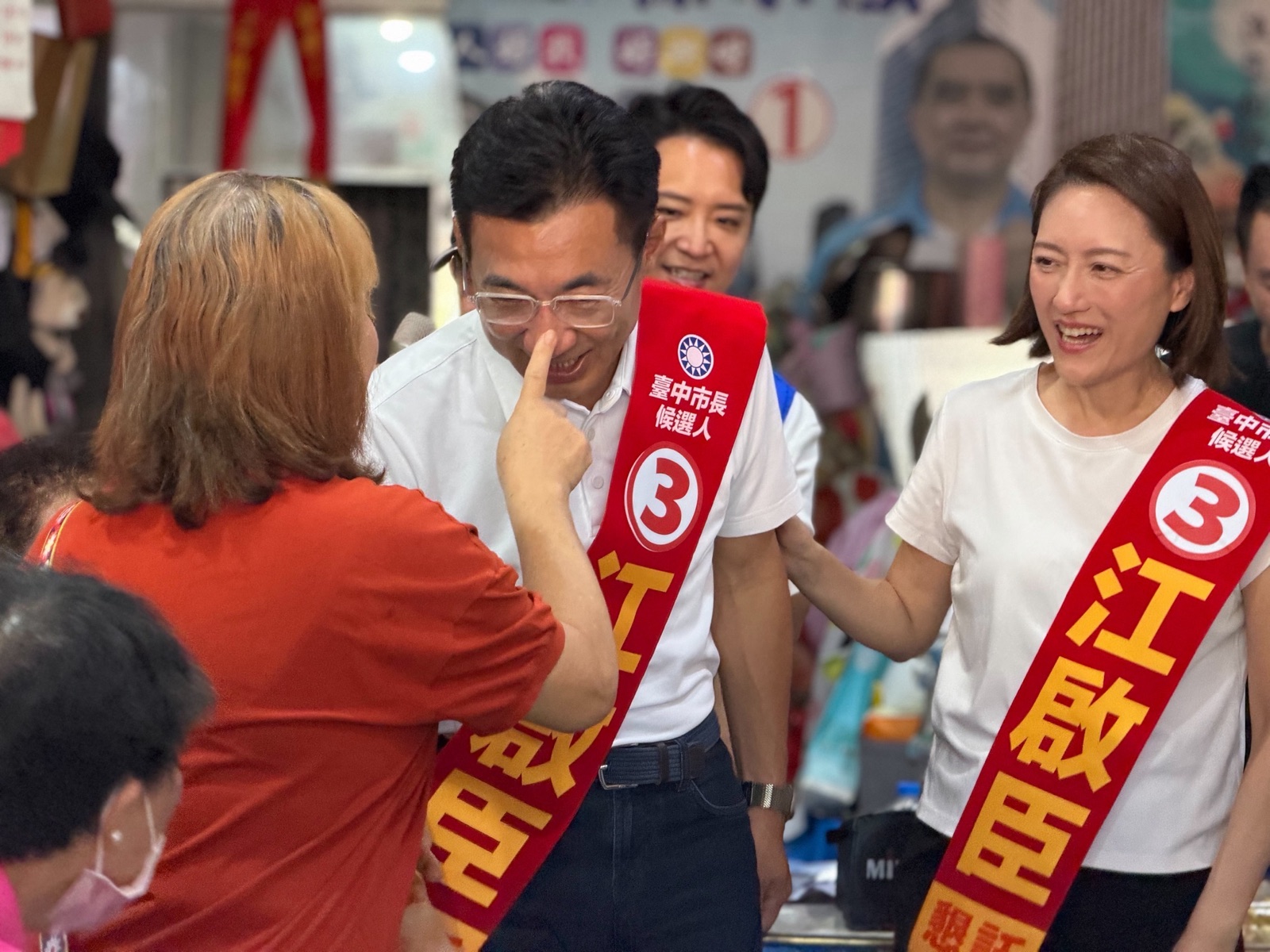 市場攤位前，穿紅色上衣的攤商女士伸手點向披「3 江啟臣」紅色肩帶男子的鼻子，男子笑著低頭，右側披同款肩帶的女士也笑著看向他