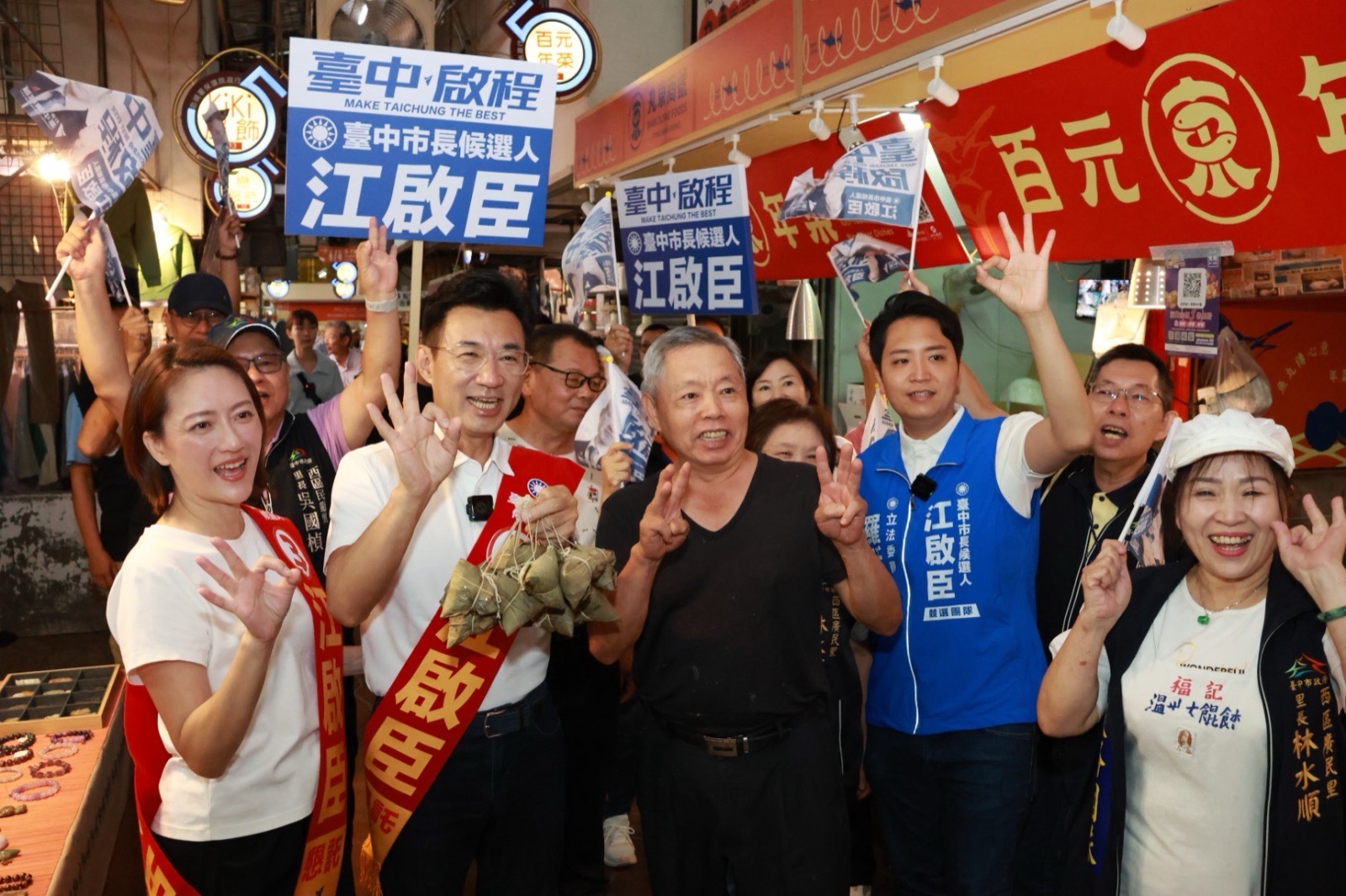 第五市場內，穿白色上衣、披「江啟臣」肩帶的男子手提一串粽子，與披同款肩帶的女士、攤商及隨行人員一同比出「OK」的3號手勢，後方舉著「臺中啟程 江啟臣」看板，右側里長背心寫著「西區廣民里 里長 林水順」