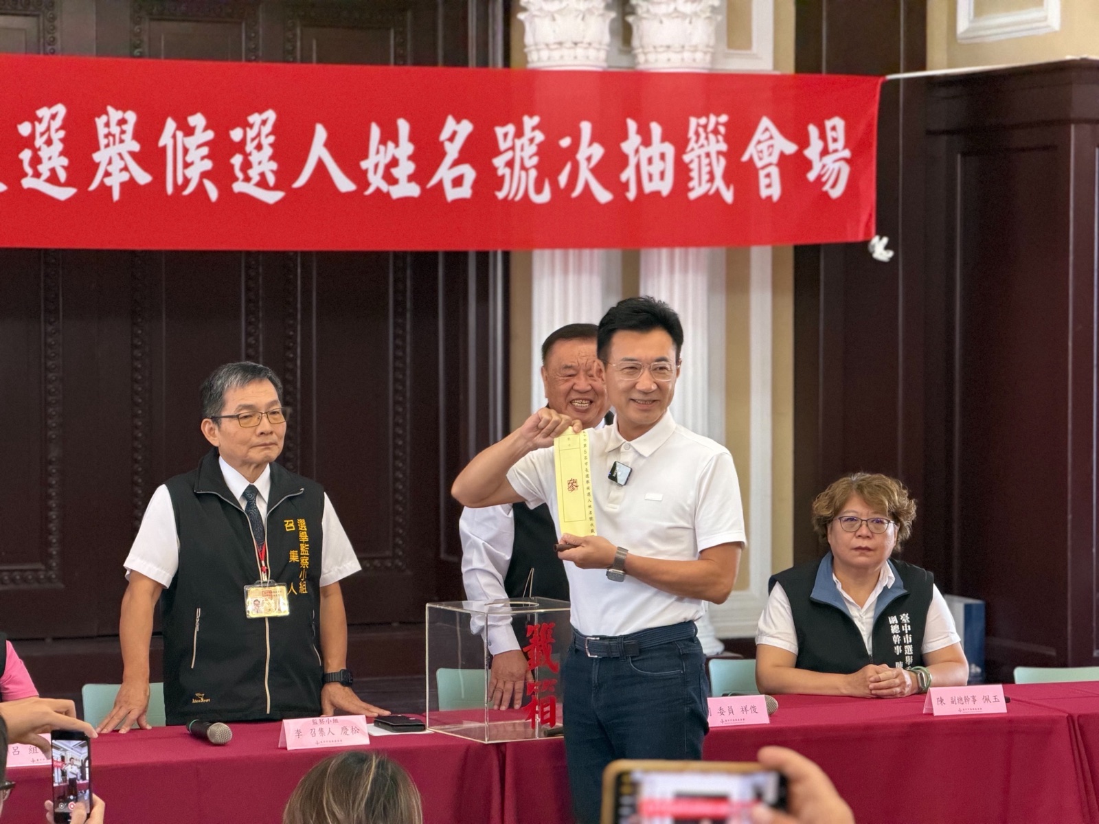 市長候選人姓名號次抽籤會場，穿白色Polo衫的男子在寫著「抽籤會場」的紅布條與透明抽籤箱前，高舉剛抽出的黃色籤條，兩側站著穿「選舉監察小組」「臺中市選舉」背心的工作人員