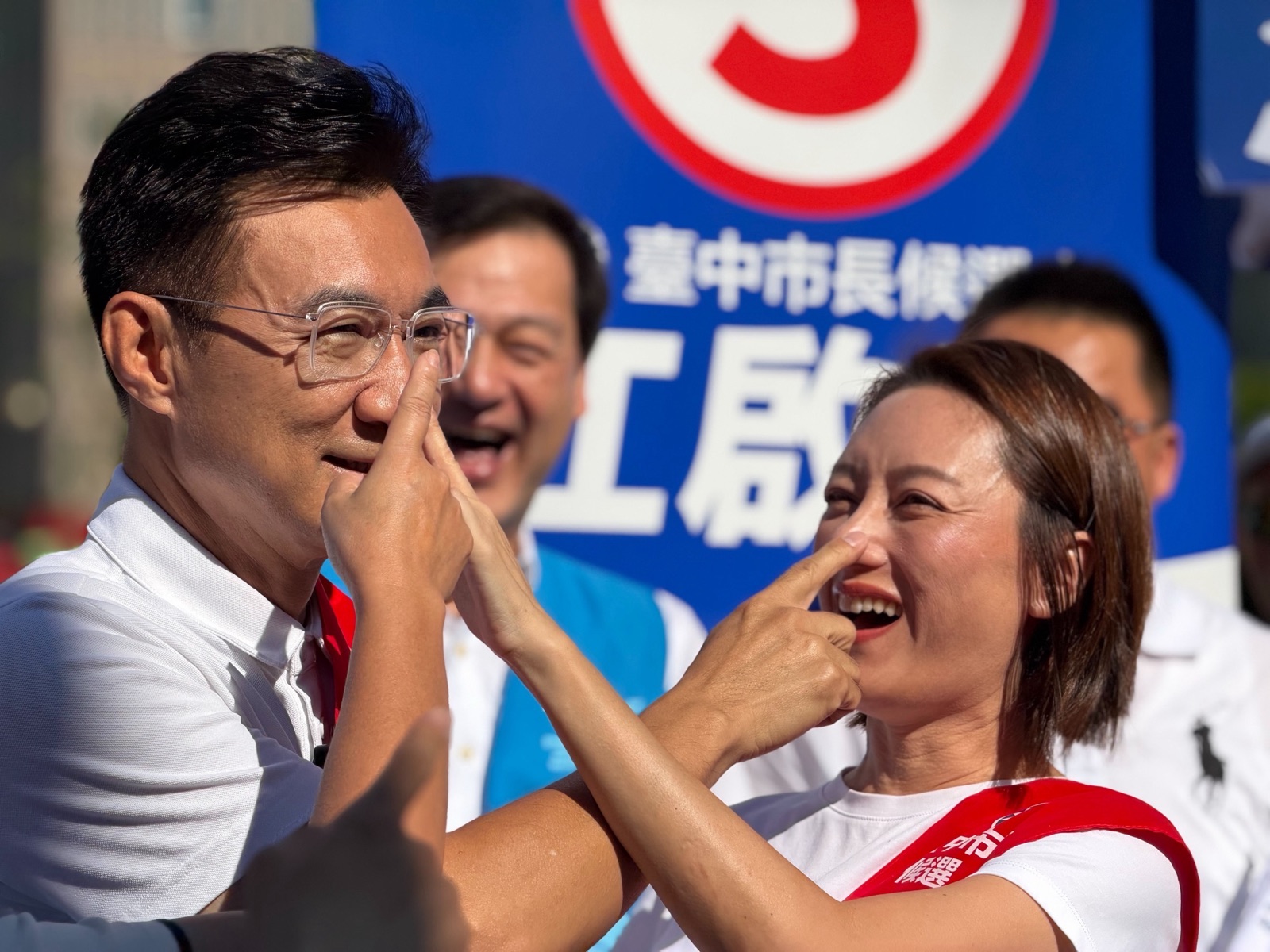 穿白色上衣的男子與身旁披「臺中市長候選人」紅色肩帶的女士，互相伸手輕點對方的鼻子，兩人開懷大笑，背後是寫有「3」與「臺中市長候選人 江啟臣」的藍色看板