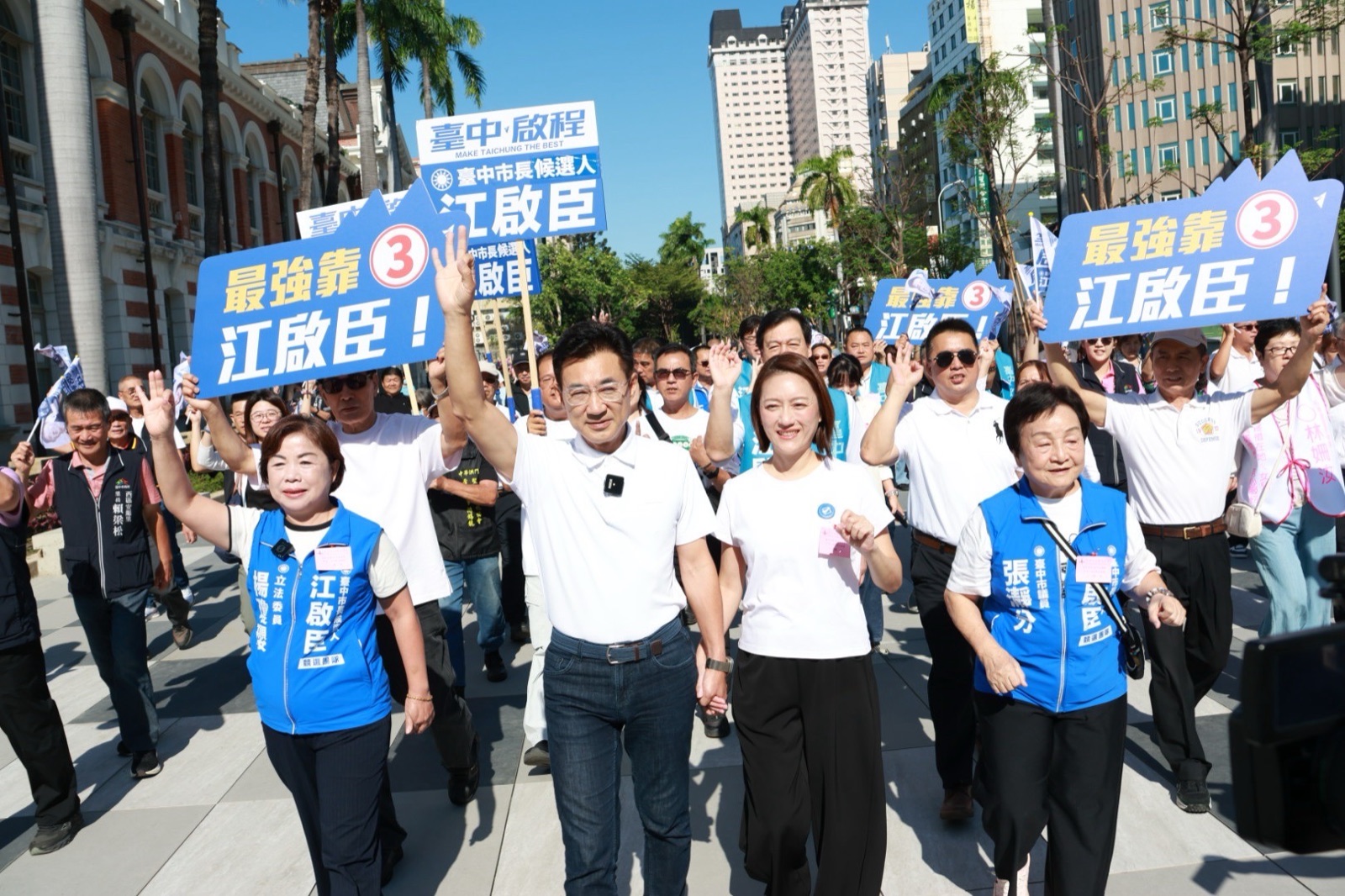徒步隊伍走在州廳前的廣場上，穿白色Polo衫的男子與身旁穿白色上衣的女士高舉三指手勢，眾人舉著「最強靠 3 江啟臣！」與「臺中啟程 臺中市長候選人 江啟臣」看板，左側穿藍背心的女士背心寫著「立法委員」，右側背心寫著「臺中市議員 張瀞分」