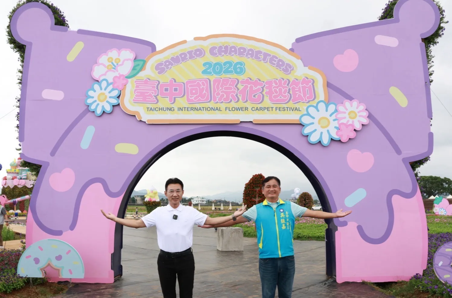 穿白色Polo衫的男子與穿淺藍色「臺中市議員 吳振嘉」背心的男子在紫色蝴蝶結造型拱門前張開雙臂合影，拱門上寫著「2026 臺中國際花毯節」