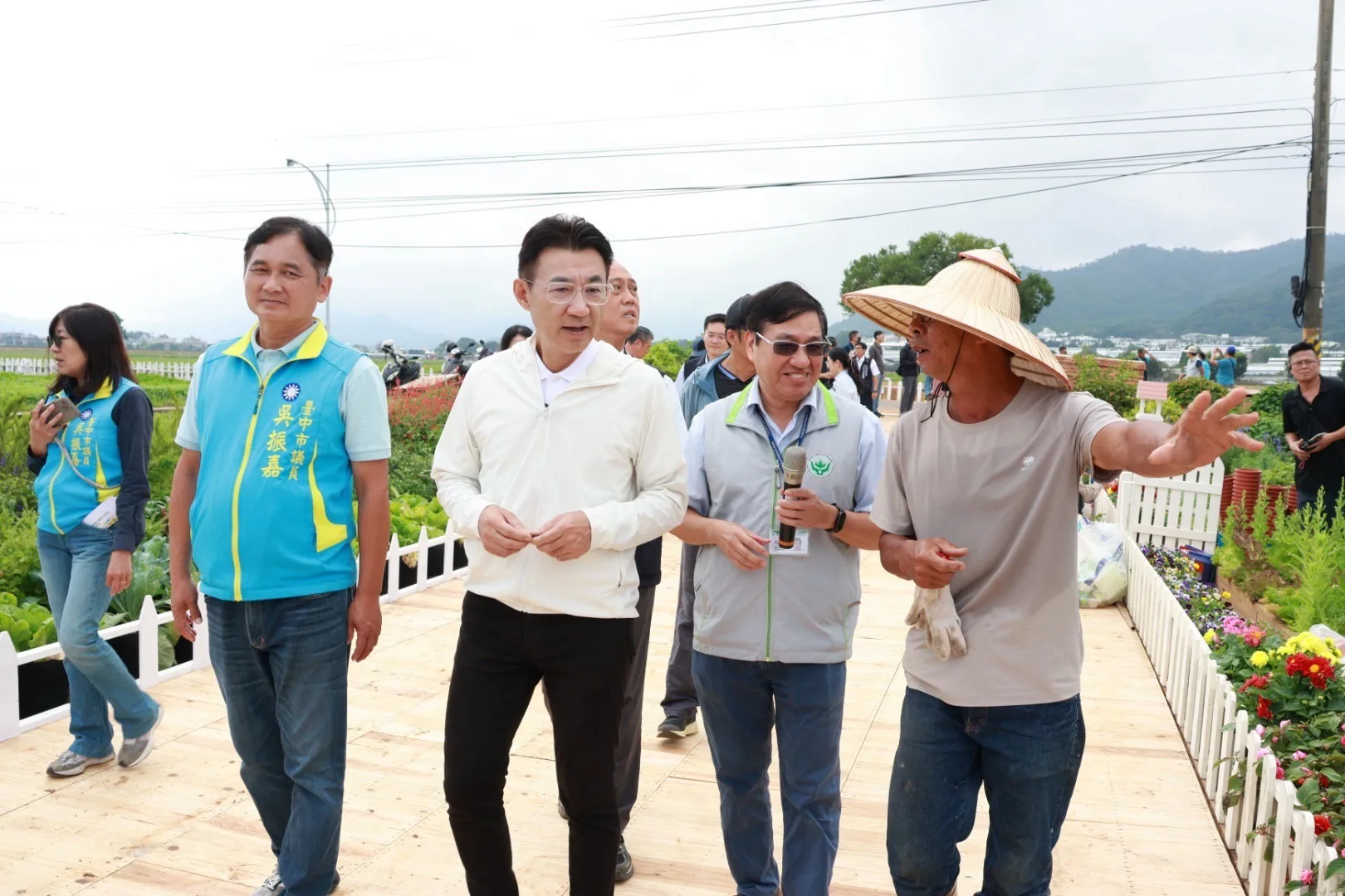 花海園區的木棧道上，穿米白外套的男子與穿淺藍色「臺中市議員 吳振嘉」背心的男子並肩行走，右側戴斗笠的農民伸手比向遠處，旁邊手持麥克風、穿灰色背心的男子面帶笑容，背景是遠山與花圃