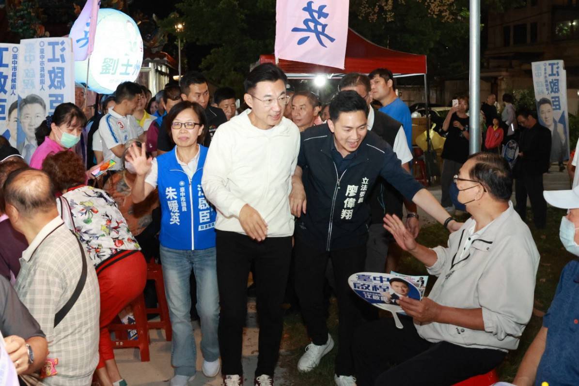 穿白色外套的男子與穿深色「立法委員 廖偉翔」外套的男子走進坐滿鄉親的廟前空地，與坐著的民眾握手，左側穿藍背心的女士背心寫著「朱暖英」