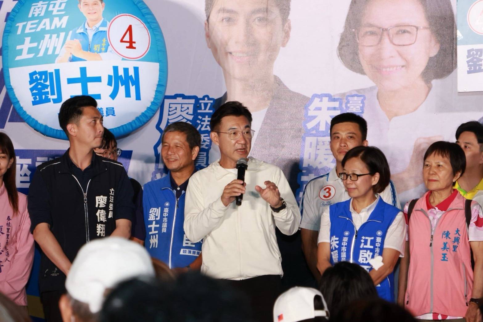 穿白色外套的男子手持麥克風發言，兩側站著穿「立法委員 廖偉翔」深色外套的男子，以及背心寫著「劉士州」「朱暖英」的人，右側穿粉紅背心者寫著「大興里 陳美玉」