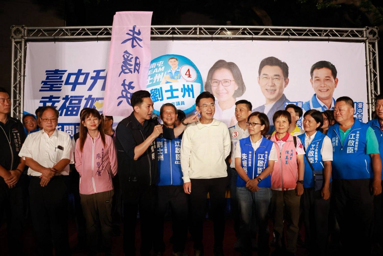 穿白色外套的男子站在臺上，身旁穿深色外套的男子手持麥克風，伸手指向他的臉頰，兩側穿藍色背心的人背心寫著「劉士州」「朱暖英」，背板寫著「臺中升級 幸福加給」