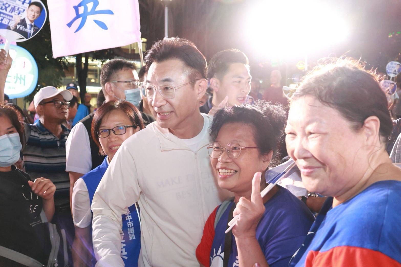 夜間廟前，穿白色外套的男子與身旁的鄉親女士合影，女士比出讚的手勢，後方穿藍背心者背心寫著「臺中市議員」，旗幟與扇子印有「江啟臣」
