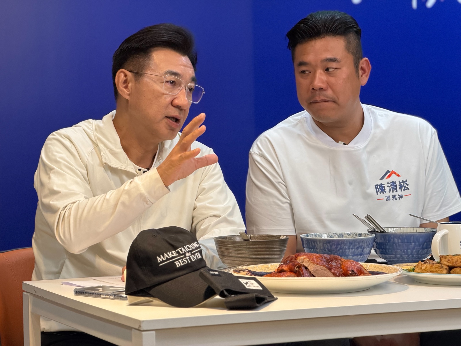 穿白色連帽外套的男子一邊比著手勢說明，一邊面向桌上的美食，身旁穿「陳清崧 潭雅神」白T的男子轉頭聆聽，桌上放著「MAKE TAICHUNG the BEST EVER」黑色帽子