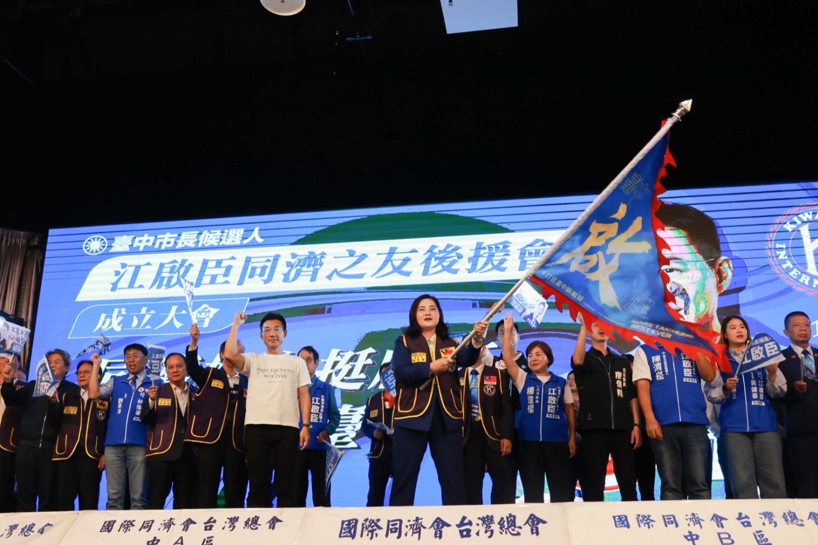 「江啟臣同濟之友後援會成立大會」舞臺上，穿紫色同濟會背心的女士揮舞藍底「啟」字戰旗，穿白T的男子與嘉賓握拳高舉，背後大螢幕寫著「臺中市長候選人 江啟臣同濟之友後援會 成立大會」