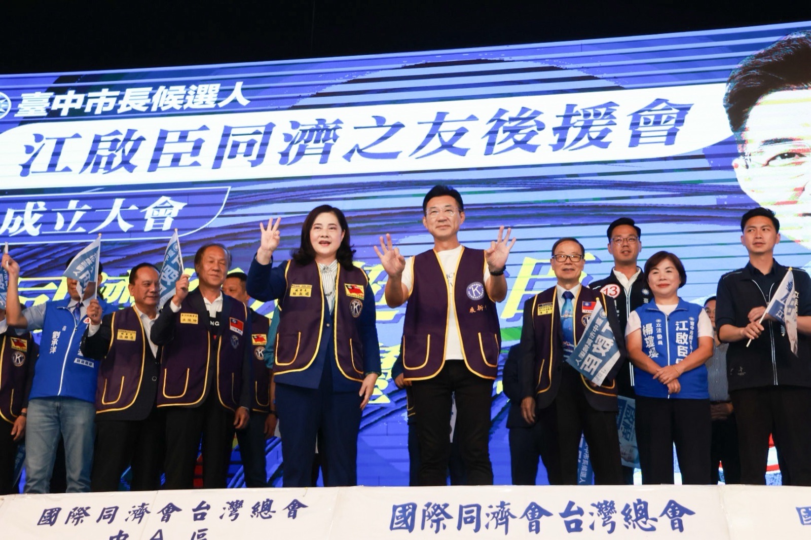 舞臺上，穿紫色同濟會背心的女士與穿紫色背心、白色上衣的男子一同比出三指手勢，旁邊站著穿藍背心的「立法委員 楊瓊瓔」，背後螢幕寫著「江啟臣同濟之友後援會」
