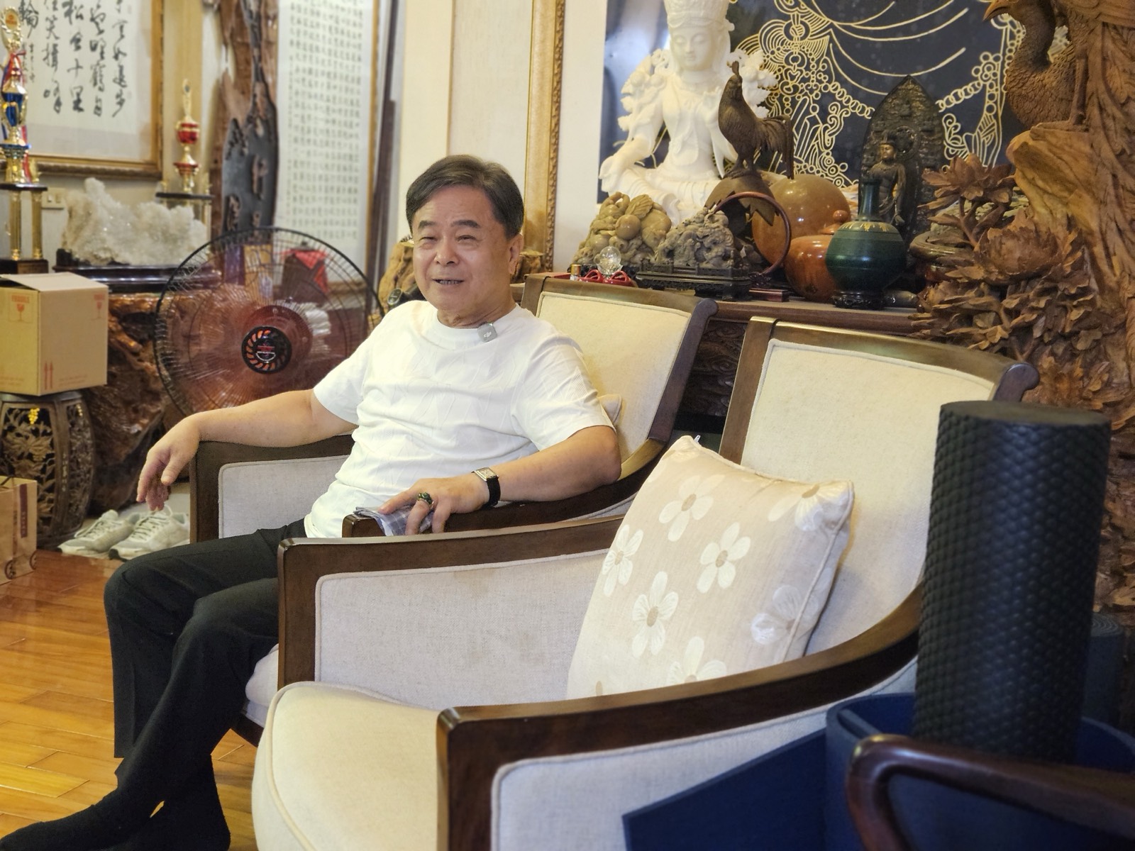 周良敦坐在工作室的扶手椅上微笑，身後擺滿佛像、木雕與書法作品