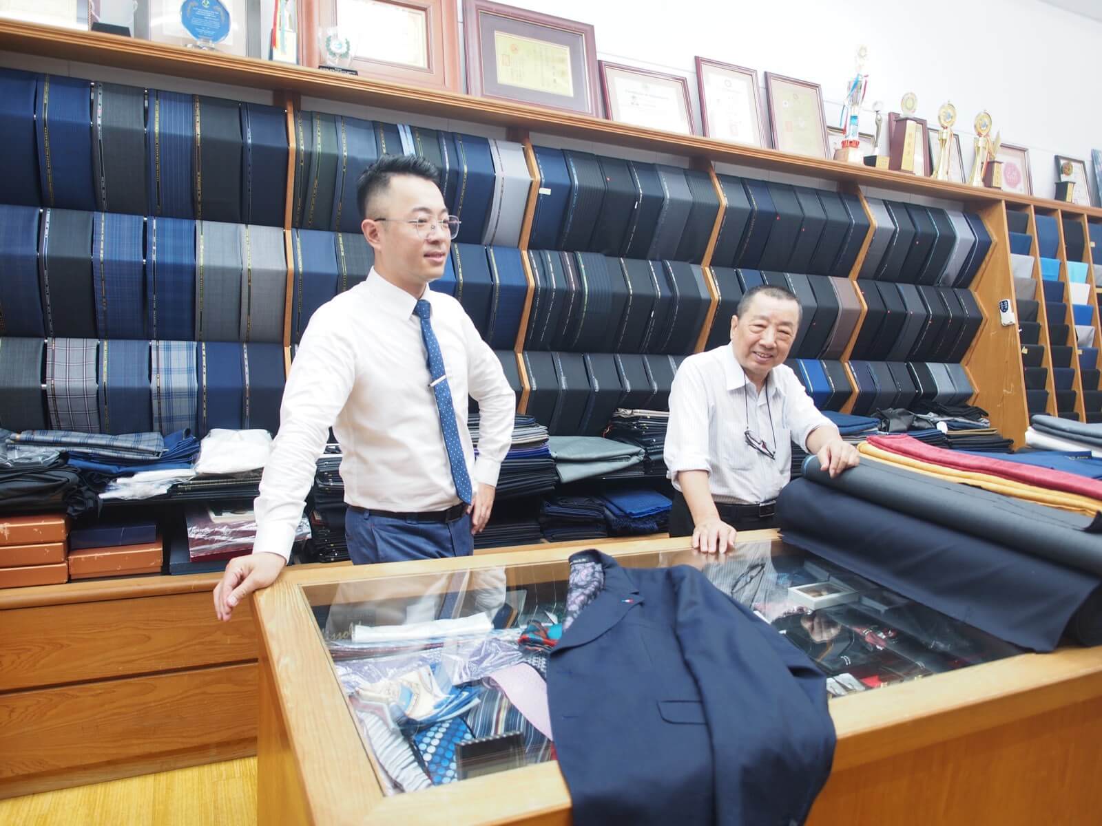 利士杰與利文雄一同站在店內的展示櫃前，櫃上攤著一件深藍色訂製西裝